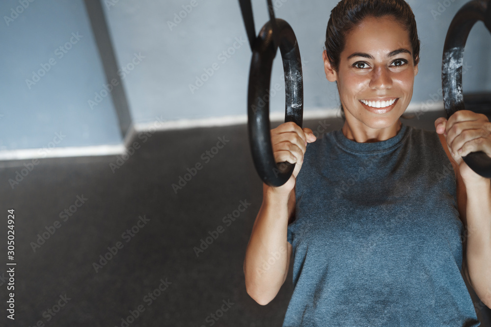 Upper-angle shot smiling sportswoman, female athlete in sportswear pumping biceps, push-up using gym equipment, hanging on rings and grinning, look camera, relaxed with sweat on forehead Upper-angle shot smiling sportswoman, female athlete in sportswear pumping biceps, push-up using gym equipment, hanging on rings and grinning, look camera, relaxed with sweat on forehead