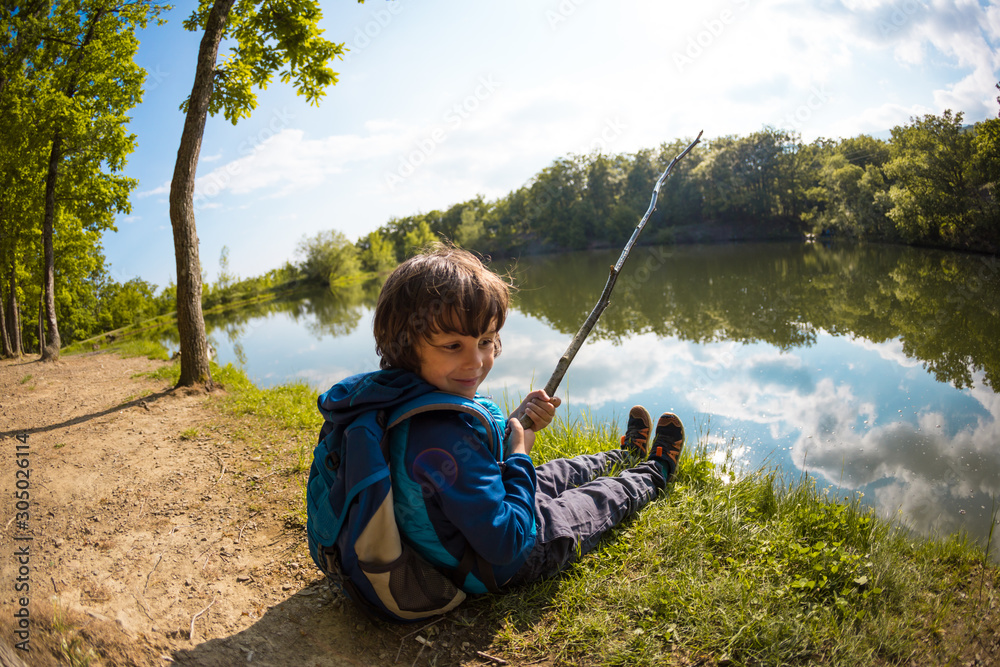 A boy with a backpack sits on the lake with a wooden stick. A boy with a backpack sits on the lake with a wooden stick.
