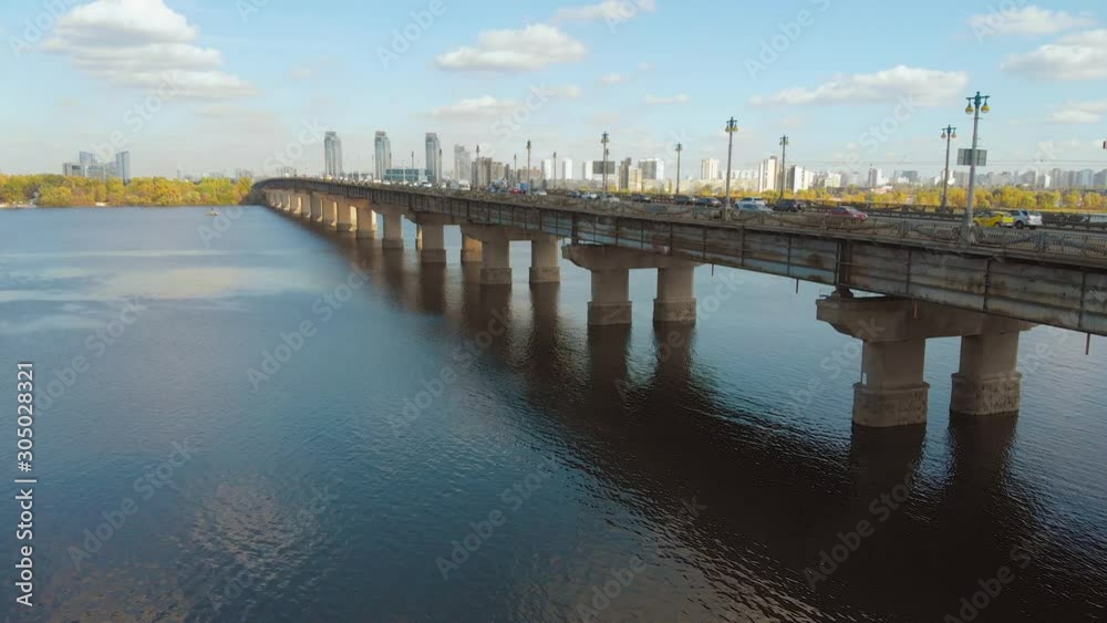 View of the city of Kiev across the Dnieper River, with a long bridge in the foreground. Cars move on the bridge in both directions. Traffic on a bridge over a wide river in the autumn. View of the city of Kiev across the Dnieper River, with a long bridge in the foreground. Cars move on the bridge in both directions. Traffic on a bridge over a wide river in the autumn.