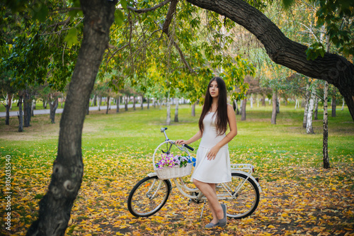 A girl in a white skirt and a vest with a bicycle and a basket of flowers stands on yellow leaves in the park in the evening, in the autumn.