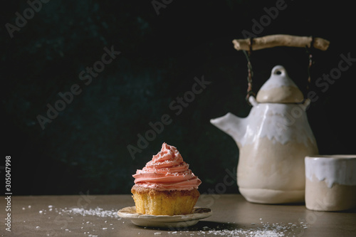Homemade cupcake with pink buttercream and coconut flakes served with ceramic teapot, cup of tea on dark table. Copy space.