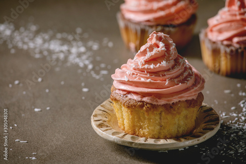 Homemade cupcakes with pink buttercream and coconut flakes served on ceramic plate on dark table. Close up