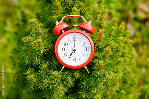 round red classic alarm clock with time at 07:00 is placed in the fir branches of a small christmas tree.