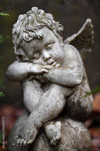 A weathered sandstone sculpture of a sad looking angel with spread wings and crossed legs sits on a globe. © Frank Middendorf
