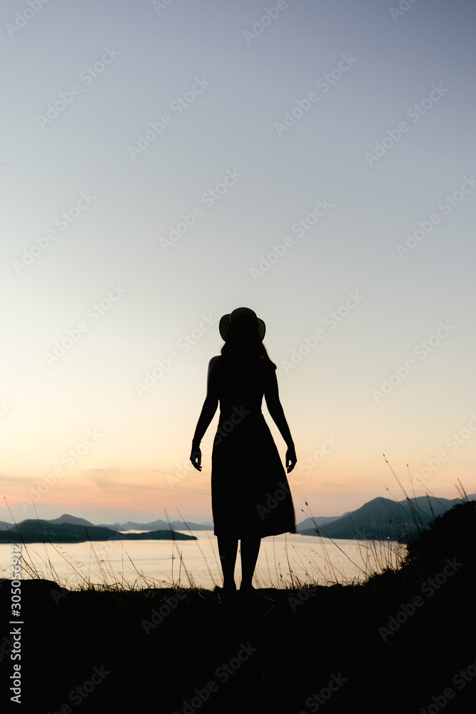 Fototapeta premium Silhouette of a woman from her back standing on a mountain at sunset in orange hues. The concept of confidence, peace and thoughts about the future