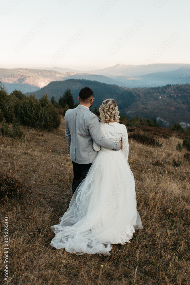 Beautiful couple having a romantic moment on their weeding day, in mountains at sunset. Bride is in a white wedding dress with a bouquet of sunflowers in hand, groom in a suit. Walking together.