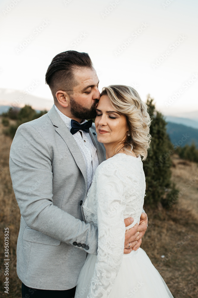 Beautiful couple having a romantic moment on their weeding day, in mountains at sunset. Bride is in a white wedding dress with a bouquet of sunflowers in hand,groom in a suit.He kissed her in forehead Beautiful couple having a romantic moment on their weeding day, in mountains at sunset. Bride is in a white wedding dress with a bouquet of sunflowers in hand,groom in a suit.He kissed her in forehead