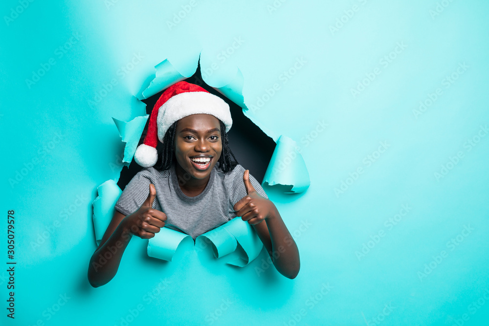 Young african woman wear in santa hat with thumbs up standing in green torn paper hole background Young african woman wear in santa hat with thumbs up standing in green torn paper hole background