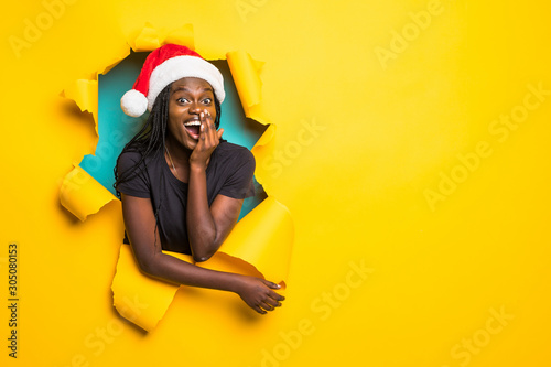 Young charming african surprised woman wear in santa hat standing in yellow torn paper hole background © F8  \ Suport Ukraine
