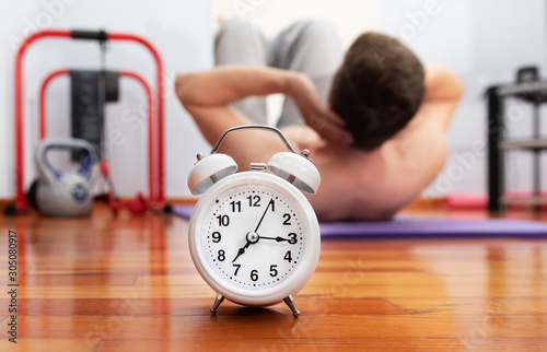 White classic alarm clock on wooden floor and unfocused blurred man exersising abs on background. 10 minutes home workout routine concept. photo