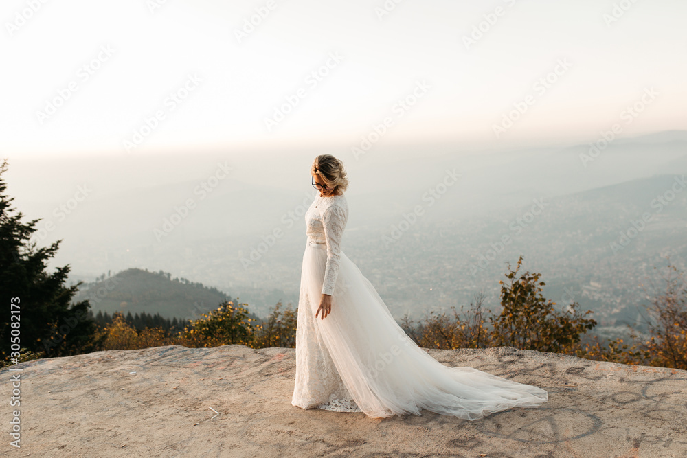 Beautiful elegant bride in lace wedding dress with long full skirt and long sleeves. Pretty girl in white. Nature, with city in the background, sunset time. Beautiful elegant bride in lace wedding dress with long full skirt and long sleeves. Pretty girl in white. Nature, with city in the background, sunset time.
