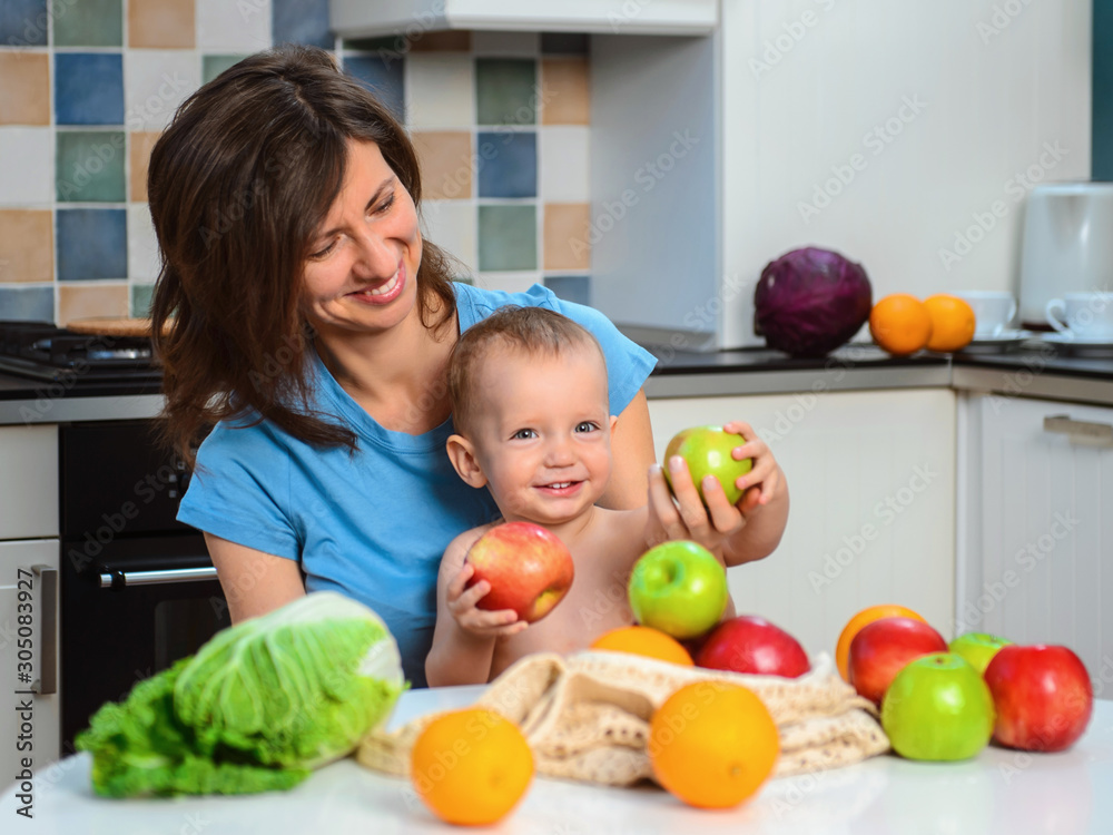 Fototapeta premium Happy mother and son baby boy eat healthy vegetarian food, lots of fruit and vegetables. Healthy nutrition for children. Fruits in ecological bag.