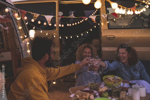 Happy friends cheering and drinking wine during camping vacation at the beach with vintage van. People having fun at weekend summer evening with camper. Youth lifestyle togetherness and party concept #305087335