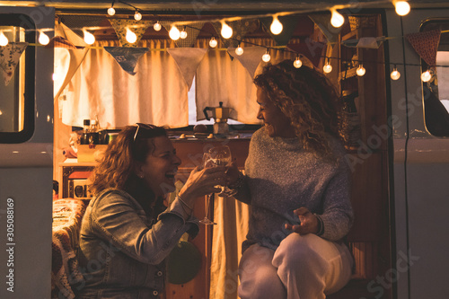 Happy couple of friends cheering and drinking wine during camping vacation in a vintage van. People having fun at weekend summer evening with camper. Youth lifestyle togetherness and party concept