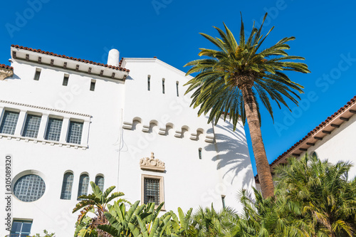 A palm tree and a white Spanish architecture style building with ornate windows and trim under a beautiful blue sky