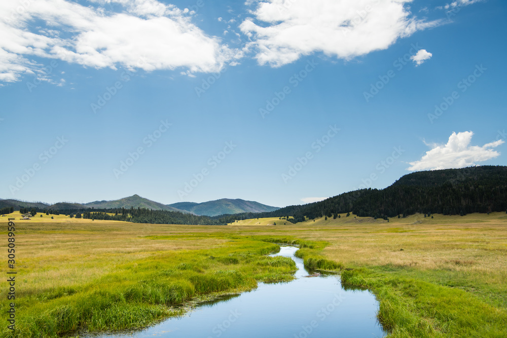 A calm stream reflects a beautiful blue sky as it curves through lush green meadows in the mountains of northern New Mexico