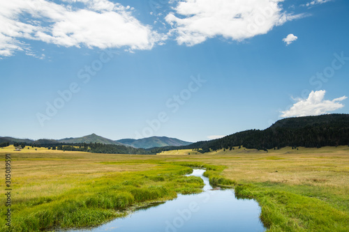 A calm stream reflects a beautiful blue sky as it curves through lush green meadows in the mountains of northern New Mexico