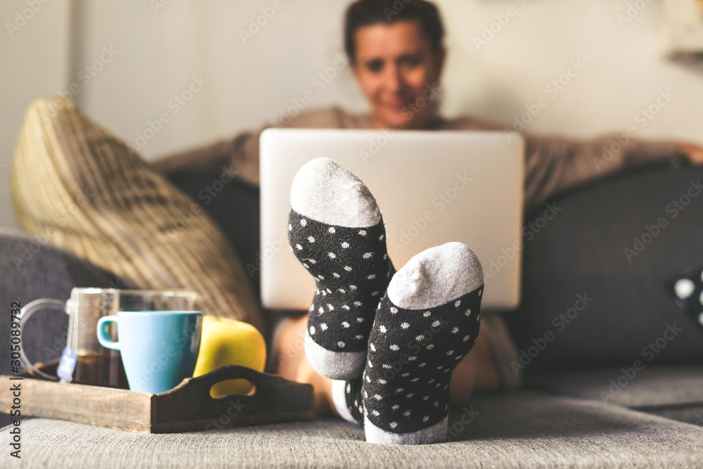 Woman sitting on a couch in the living room with warm socks in a winter morning. Girl using laptop and works at home, having natural breakfast with tea coffee and fruits. Focus on feet in foreground - obrazy, fototapety, plakaty 