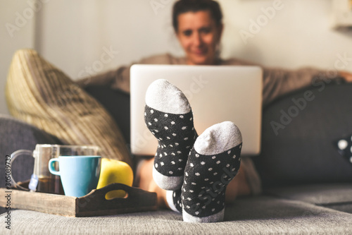 Woman sitting on a couch in the living room with warm socks in a winter morning. Girl using laptop and works at home, having natural breakfast with tea coffee and fruits. Focus on feet in foreground