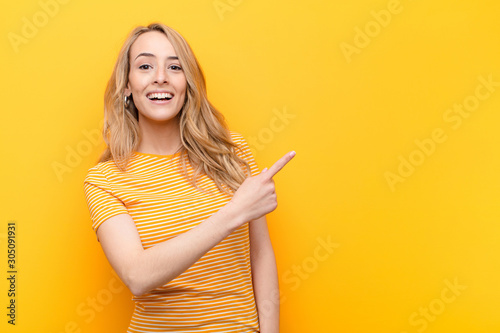 young pretty blonde woman looking excited and surprised pointing to the side and upwards to copy space against flat color wall photo