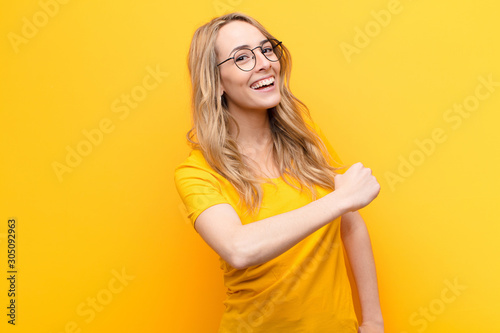 young pretty blonde woman feeling happy, positive and successful, motivated when facing a challenge or celebrating good results against flat color wall