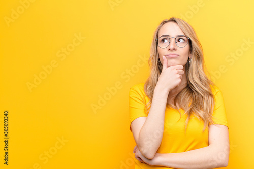 young pretty blonde woman thinking, feeling doubtful and confused, with different options, wondering which decision to make against flat color wall