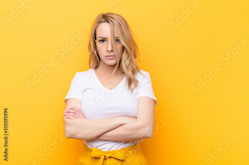 young pretty blonde woman feeling displeased and disappointed, looking serious, annoyed and angry with crossed arms against flat color wall