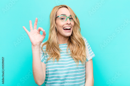 young pretty blonde woman feeling successful and satisfied, smiling with mouth wide open, making okay sign with hand against flat color wall