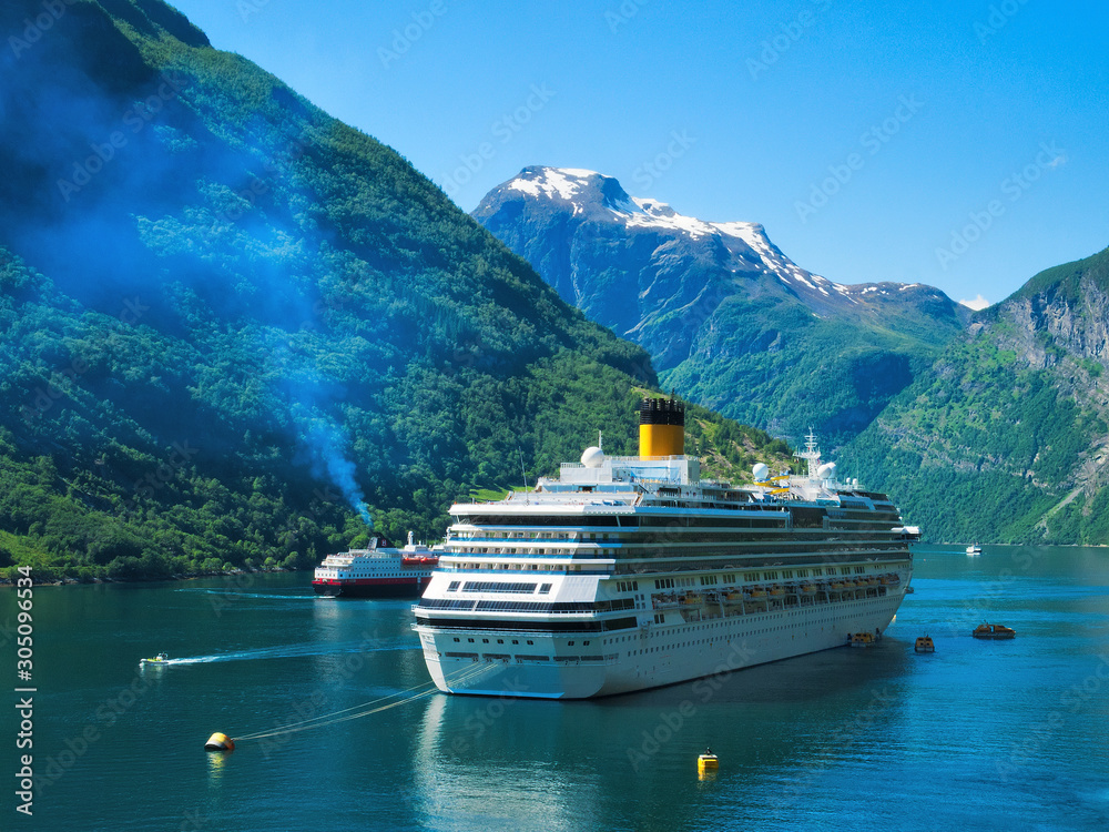 Gorgeous view of Geiranger Fjord and surrounding mountains with snow, waterfalls, moored cruise ship and tender boats carrying tourists from ship to shore on a beautiful day with blue sky. Gorgeous view of Geiranger Fjord and surrounding mountains with snow, waterfalls, moored cruise ship and tender boats carrying tourists from ship to shore on a beautiful day with blue sky.