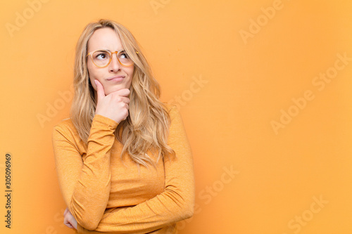 young pretty blonde woman thinking, feeling doubtful and confused, with different options, wondering which decision to make against flat color wall