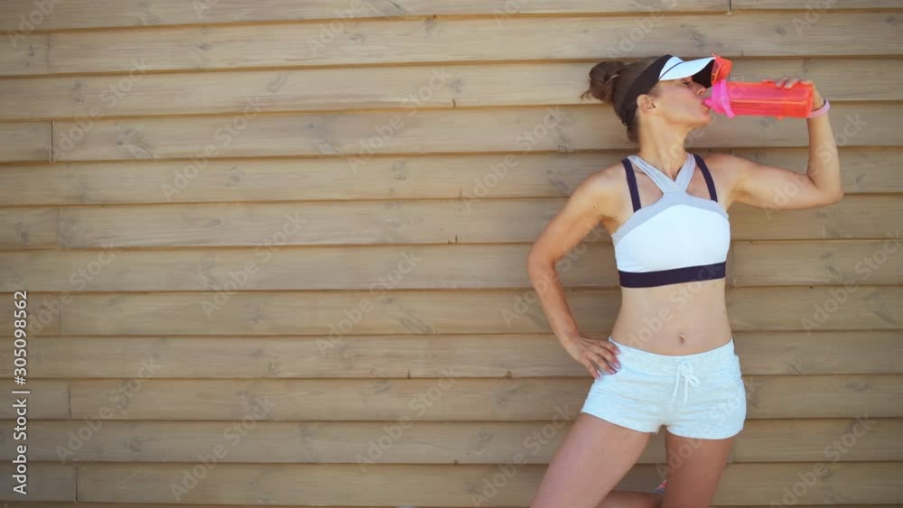 young woman jogger in fitness clothes drinking water from bottle against wooden wall.