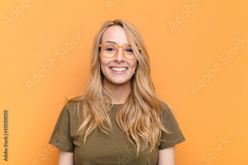young pretty blonde woman looking happy and pleasantly surprised, excited with a fascinated and shocked expression against flat color wall