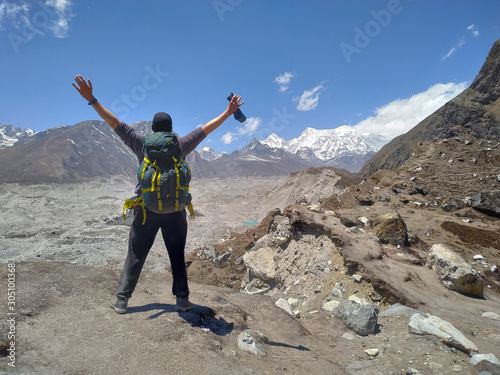 Traveler with green backpack stands with open arms in front of Ngozumpa glacier covered with stones in Himalayas in Nepal. Snowy Cho Oyu mountain is visible in the background.