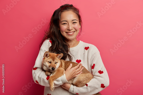 Photo of positive Asian girl gets cute shiba inu puppy as present, ready to care about domestic pet, likes playing with animals, spend day together. Dog walker takes pup for walk, isolated on pink