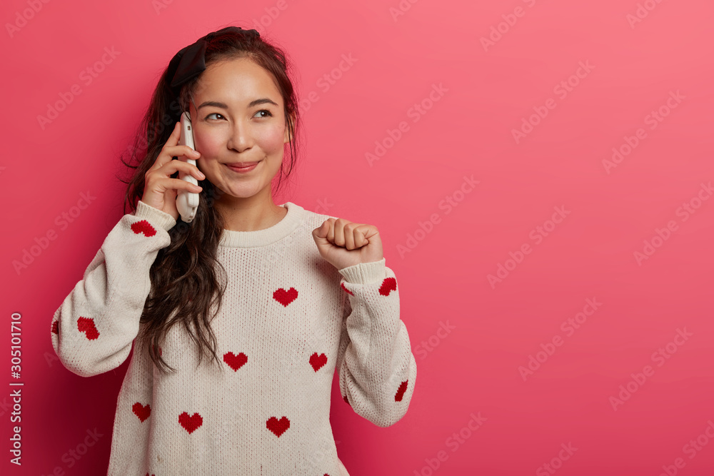 Joyful girlfriend enjoys casual friendly conversation with pal, talks and rejoices cool news, looks aside with dreamy expression, raises clenched fist, keeps smartphone pressed to ear. Technology