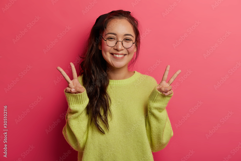 Lucky cheerful girl shows victory or peace gesture, enjoys awesome day, sends positive vibes, has toothy smile, dark hair combed in pony tail, dressed in green jumper, models over pink background Lucky cheerful girl shows victory or peace gesture, enjoys awesome day, sends positive vibes, has toothy smile, dark hair combed in pony tail, dressed in green jumper, models over pink background