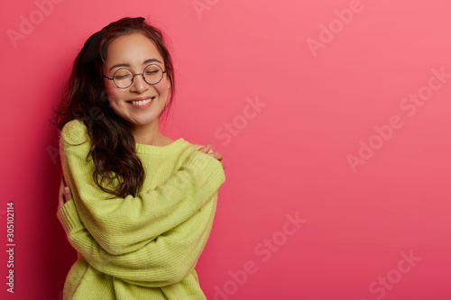 Photo of charming woman expresses self love and care, enjoys wearing new soft green sweater, embraces own body, recalls romantic moment with boyfriend, smiles pleasantly, stands over rosy wall photo