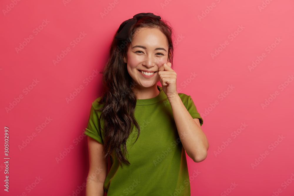 Beautiful young natural brunette girl touches cheek gently, has healthy skin, shapes mini heart with fingers, has charming appealing smile, wears green t shirt, isolated over vivid pink background. Beautiful young natural brunette girl touches cheek gently, has healthy skin, shapes mini heart with fingers, has charming appealing smile, wears green t shirt, isolated over vivid pink background.