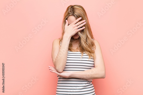 young pretty blonde woman looking stressed, ashamed or upset, with a headache, covering face with hand against flat color wall