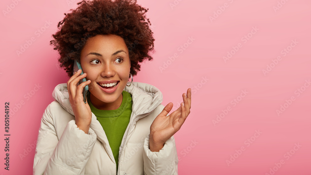 Studio shot of smiling glad adult woman has telephone conversation, keeps palm raised, dressed in winter coat, discusses plans for weekend, uses mobile phone, isolated on pink wall, blank space Studio shot of smiling glad adult woman has telephone conversation, keeps palm raised, dressed in winter coat, discusses plans for weekend, uses mobile phone, isolated on pink wall, blank space