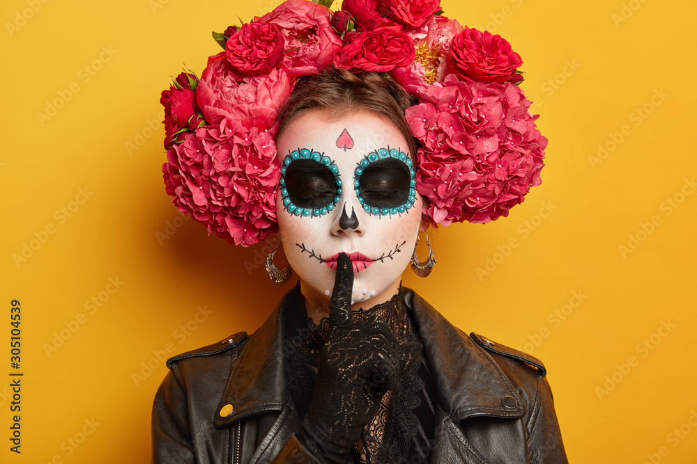 Close up shot of secret woman has sugar skull makeup makes silence gesture keeps finger over lips, stands with closed eyes dark painted circles around being charming and dangerous. Day of Dead concept Close up shot of secret woman has sugar skull makeup makes silence gesture keeps finger over lips, stands with closed eyes dark painted circles around being charming and dangerous. Day of Dead concept