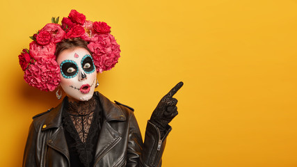 Shocked female wears creative sugar skull makeup, wreath made of red peonies, celebrates All Souls day, holiday in Mexico points away with index finger isolated over yellow background shows copy space