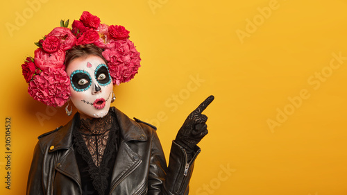 Shocked female wears creative sugar skull makeup, wreath made of red peonies, celebrates All Souls day, holiday in Mexico points away with index finger isolated over yellow background shows copy space