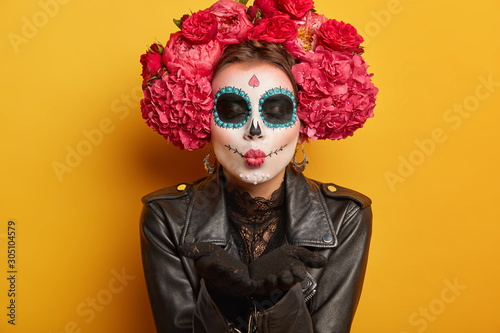 Photo of lovely woman blows mwah, keeps lips folded, wears creative makeup, prepares for carnival, prepares for Day of Dead, expresses love, poses against yellow background. Mexican female sends kiss