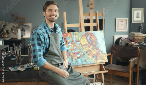 Portrait of contented young male painter sitting on a background of his new painting