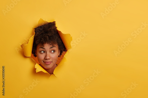 Photo of attractive curly African American woman looks with curious expression aside, notices something interesting, has natural beauty, isolated over yellow background in paper hole, has fun indoor photo