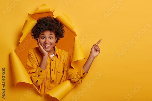 Fotografie Horizontal view of ethnic lady gives instruction how to get place, promots copy space, touches cheek with palm, dressed in fashionable clothing, smiles pleasantly