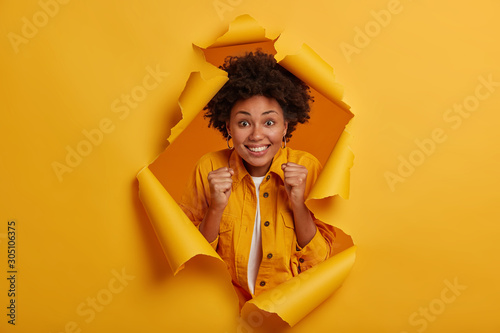 Positive lucky woman with Afro hairstyle, raises clenched fists, smiles broadly, poses in ripped hole background, enjoys perfect day, anticipates for something awesome, screams from delight. photo