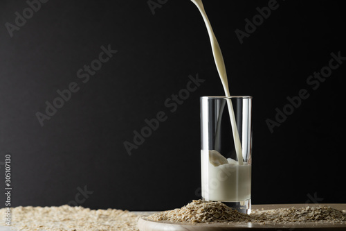 Pouring oat milk into glass on black background and oat flakes on table. Oat milk lactose free milk substitute concept. Copy space, soft shadows. photo