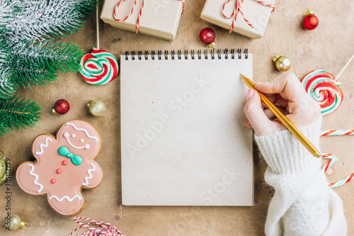 Notebook in the center of cute Christmas decoration: gift box, christmas balls, gingerbread man, fir tree branch, cande cane. Female hand writing in notebook. Flat lay. Top view photo